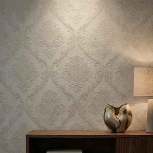 May include: Neutral-toned wallpaper with a repeating damask pattern. The design features ornate floral and scroll motifs, creating a textured, embossed effect. A wooden shelf holds a decorative vase and a lamp with a beige shade.