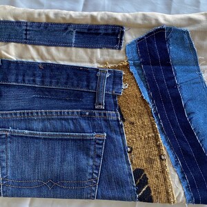 May include: A patchwork textile art piece featuring denim, beige fabric, and a woven brown material. The denim includes a back pocket and belt loops. The design incorporates various textures and shades of blue.
