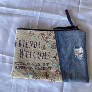 May include: A rectangular zippered pouch with a denim panel and a patterned fabric panel. The fabric panel features the text "FRIENDS WELCOME RELATIVES BY APPOINTMENT" in purple, with spiral and dot designs. The denim panel has a distressed area.