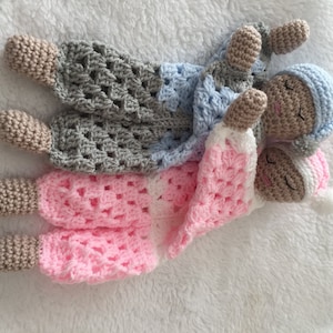May include: Three handmade crochet baby dolls in pastel shades. The dolls have closed eyes and are wearing hats. The blankets are pink, white, grey, and blue. The dolls are lying on a white, fluffy surface.