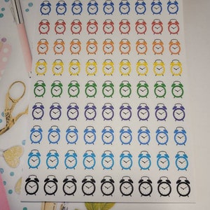 May include: Sheet of colorful alarm clock stickers in rows. The clocks are in shades of blue, red, orange, yellow, green, purple, and black. The text "ORGANIZING CHAOS" is at the top. A pink pen and gold scissors are in the background.