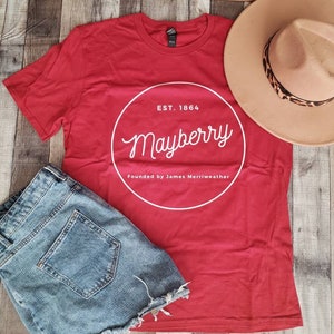 May include: Red t-shirt with a white circle graphic that says "Est. 1864 Mayberry Founded by James Merriweather".