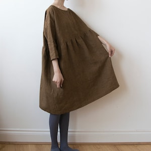 May include: A brown linen dress with three-quarter sleeves and a gathered waist. The dress is knee-length and has a loose, flowing silhouette. The model is wearing gray tights. The dress is a solid color and appears to be made of a natural fabric.