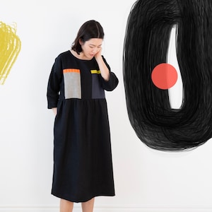 May include: A woman wearing a black dress with a geometric pattern in orange, yellow, gray, and black. The dress has a loose fit and a round neckline. The woman is standing in front of a white wall with a black and red abstract design.