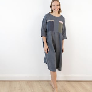 Könnte beinhalten: Eine Frau in einem grauen Leinenkleid mit Dreiviertelärmeln und einem knielangen Saum. Das Kleid hat drei rechteckige Aufnäher in verschiedenen Farben und Mustern auf dem Oberteil. Die Frau steht auf einem hellen Holzboden.
