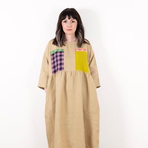 Könnte beinhalten: Ein beigefarbenes Leinenkleid mit Patchwork-Design, das aus einem grün-rosa-schwarzen karierten Stoff, einem gelben Stoff mit rosa Besatz und einem grünen Stoff mit rosa Besatz besteht.