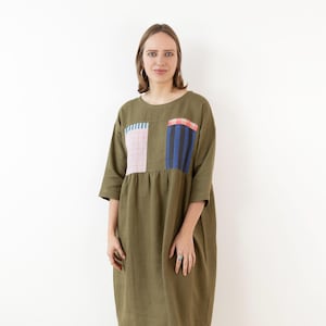 Könnte beinhalten: Olivgrünes Kleid mit Dreiviertelärmeln und zwei quadratischen Taschen. Eine Tasche hat ein rosa-weißes Gittermuster, die andere blaue und schwarze vertikale Streifen mit roten Akzenten. Das Kleid hat eine lockere Passform.