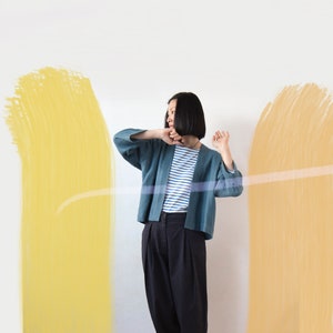 Könnte beinhalten: Eine Frau trägt ein blau-weiß gestreiftes Hemd und einen tealblauen Cardigan mit lockerer Passform. Sie steht vor einer weißen Wand mit zwei senkrechten, gelb gestrichenen Streifen.