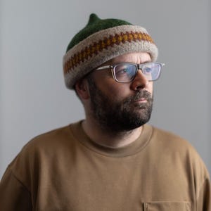 Può includere: Un berretto lavorato a maglia verde, beige e marrone, con una parte superiore a punta e un motivo geometrico marrone e giallo. La persona nell'immagine indossa una camicia marrone e occhiali.