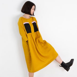 Könnte beinhalten: Ein gelbes Leinenkleid mit schwarzen und pinkfarbenen geometrischen Patches. Das Kleid hat eine lockere Passform und einen gerafften Rock. Das Model trägt schwarze Stiefeletten.