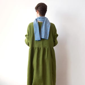 Könnte beinhalten: Eine Person trägt ein olivgrünes langes Kleid und einen blau-weiß gestreiften Schal. Das Kleid hat eine geraffte Taille und lange Ärmel. Der Schal ist um den Hals drapiert, wobei die Streifen horizontal verlaufen.