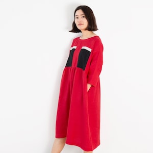 Könnte beinhalten: Ein rotes Kleid mit schwarzen und weißen Verzierungen an den Taschen. Das Kleid hat eine lockere Passform und einen runden Ausschnitt.