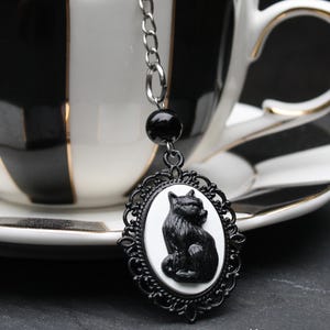 May include: A black cat tea infuser with a decorative black frame and chain, hanging in front of a black and white striped teacup. The infuser features a black cat cameo on a white background. The teacup has a gold rim.