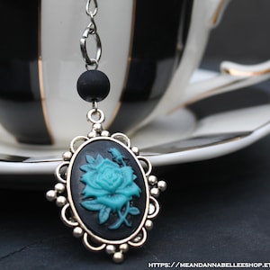 May include: A silver tea infuser with a black and teal rose cameo pendant. The pendant hangs from a chain and a black bead. The infuser is next to a black and white striped teacup with gold trim.