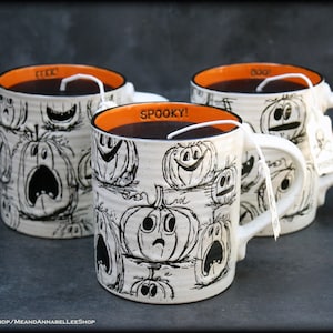 Puede incluir: Tres tazas de cerámica blanca con bordes naranjas e ilustraciones de calabazas en blanco y negro. Cada taza tiene una bolsita de té con una cuerda que cuelga de la parte superior. Las tazas están etiquetadas con "Spooky!" "Boo!" y "Trick or Treat".