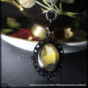 May include: A black and gold charm with a witch flying on a broomstick in a yellow and black silhouette. The charm is attached to a silver chain and a black bead.