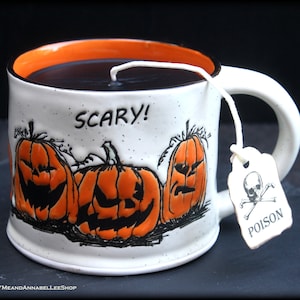 Puede incluir: Taza de Halloween de cerámica blanca con interior y asa naranja. La taza presenta tres calabazas naranjas talladas y la palabra "SCARY!" en negro. Una bolsita de té con una calavera y huesos cruzados y la palabra "POISON" cuelga del borde.