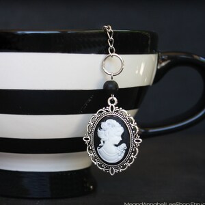 May include: A silver tea infuser charm with a black cameo featuring a woman's profile. The charm is hanging from a black and white striped teacup.