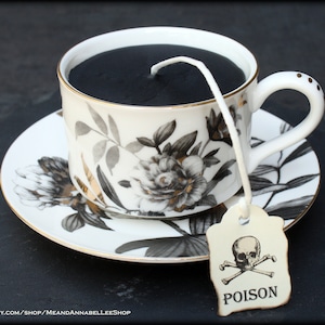 May include: A black tea candle in a white teacup with a floral pattern and gold accents. The teacup is on a matching saucer. A tag with a skull and crossbones and the word "POISON" is attached to the teacup.