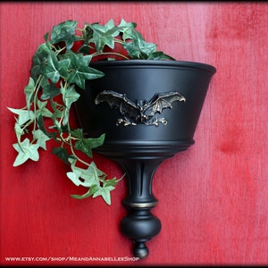 May include: Black wall-mounted planter with a bat design. The planter is filled with green ivy.