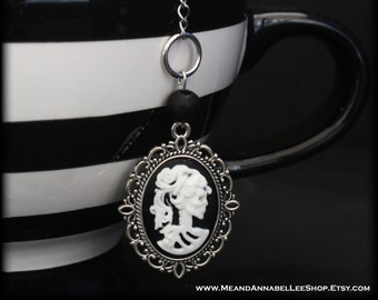 Puede incluir: Una taza de té a rayas blancas y negras con una cadena plateada y un dije de camafeo con el perfil de una mujer esqueleto blanca. El camafeo está engastado en un marco plateado con detalles ornamentados.