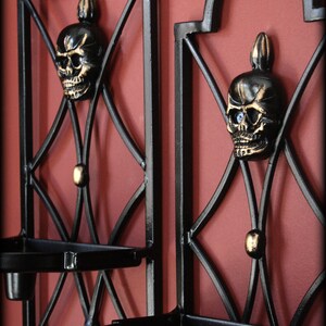 May include: Two black metal wall sconces with a skull design. Each sconce features a skull with gold accents and a small tray for holding candles. The sconces are mounted on a red wall.