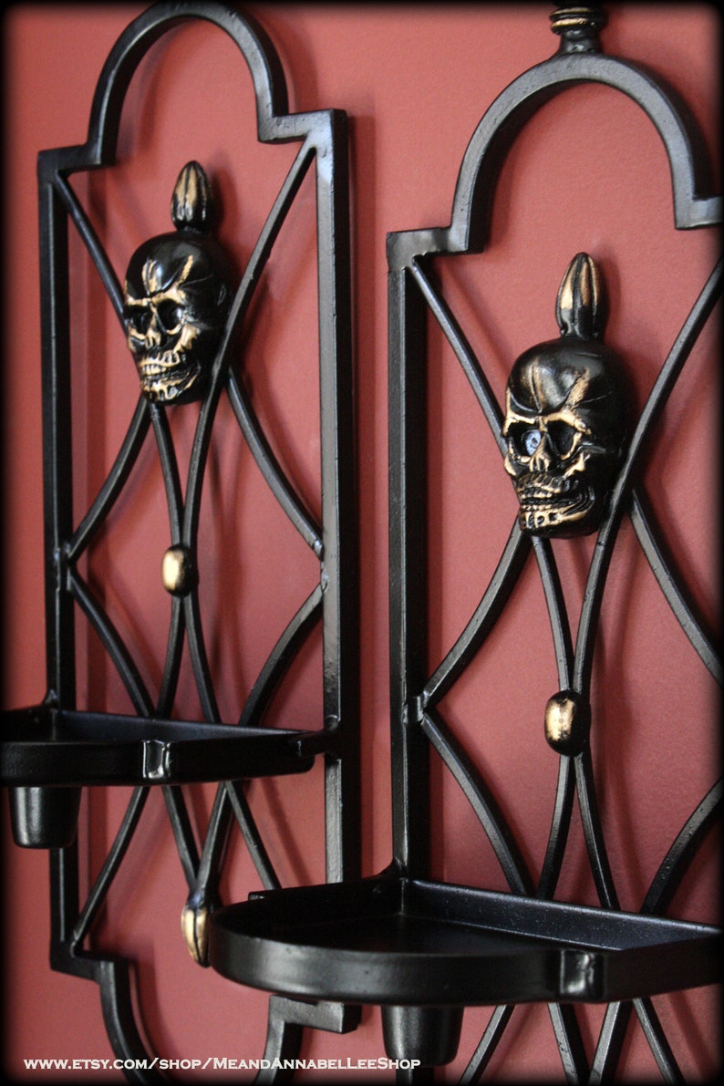 May include: Two black metal wall sconces with a skull design. Each sconce features a skull with gold accents and a small tray for holding candles. The sconces are mounted on a red wall.