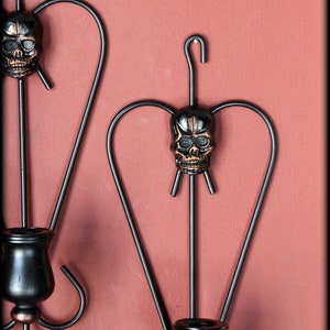 May include: Two black metal wall sconces with a skull design. Each sconce has a candle holder and a skull with a copper finish.