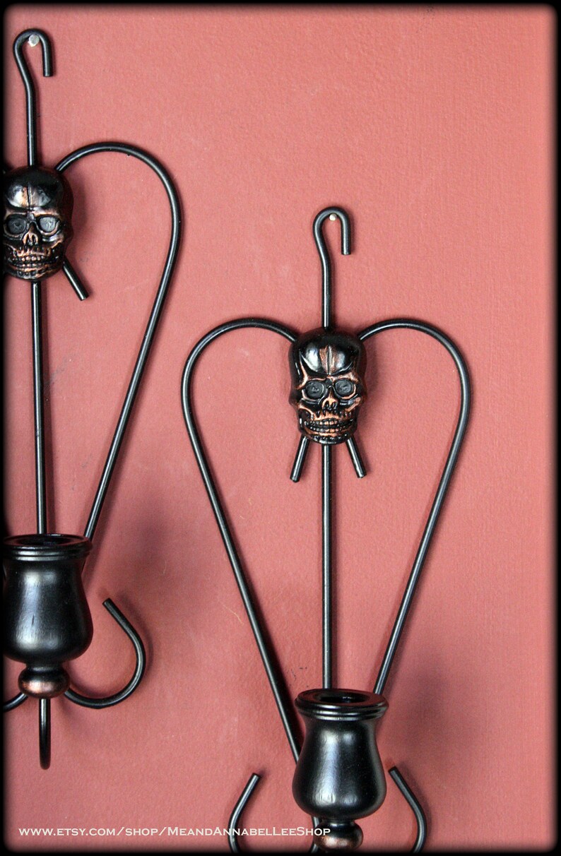 May include: Two black metal wall sconces with a skull design. Each sconce has a candle holder and a skull with a copper finish.