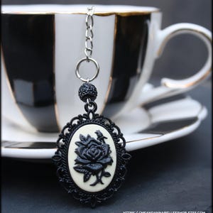 May include: Black and white tea infuser with a cameo design. The oval pendant features a black rose on a cream background, framed by a decorative black border. A small black beaded accent and a silver chain complete the design. The infuser is shown with a black and white teacup.