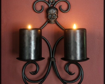 Puede incluir: Aplique de pared de metal negro con dos velas de pilar negras. El aplique presenta un diseño de calavera y adornos decorativos. Las velas están encendidas, emitiendo un brillo cálido. Una pieza única para Halloween o decoración gótica.