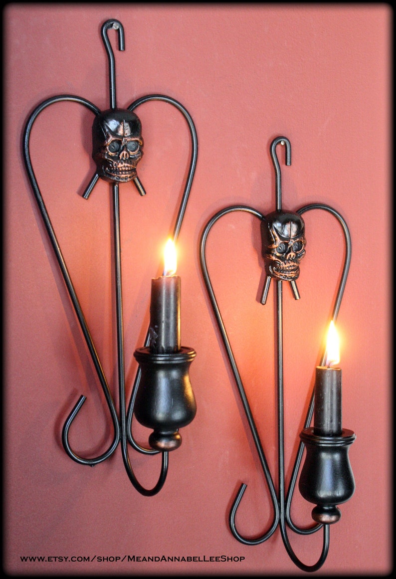 May include: Two black metal wall sconces with skull accents and candle holders. Each sconce has a heart-shaped design and a single candle holder. The sconces are mounted on a red wall.