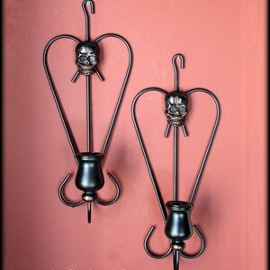 May include: Two black metal wall sconces with a heart-shaped design and a skull detail. Each sconce has a candle holder.