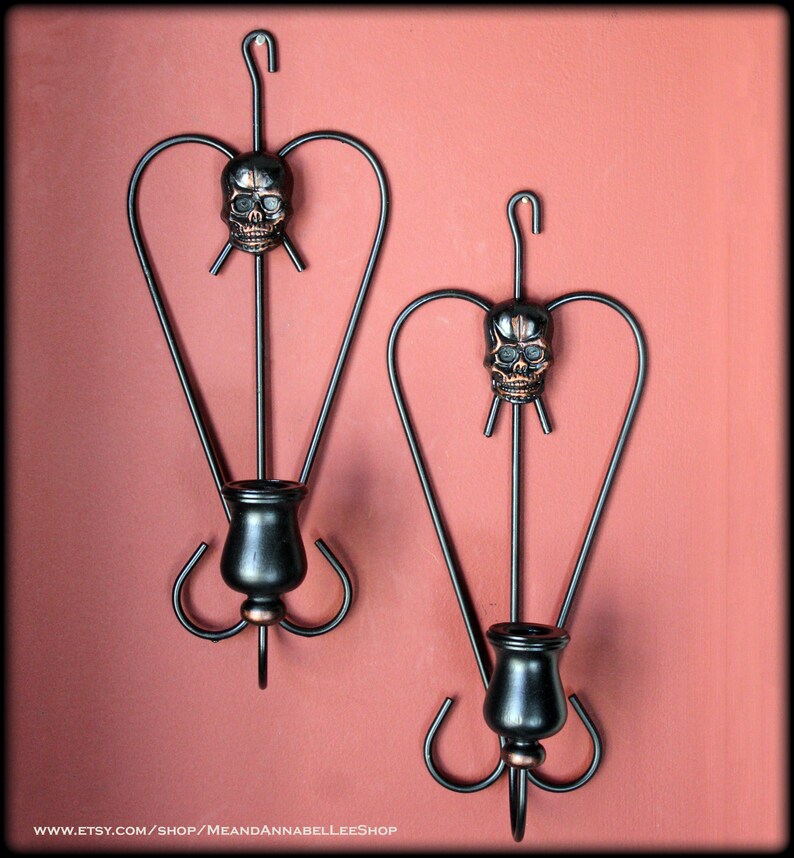 May include: Two black metal wall sconces with a heart-shaped design and a skull detail. Each sconce has a candle holder.