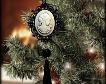 Peut inclure: Ornement de Noël avec un camée noir représentant le profil d'une femme blanche. Il est suspendu à un fil noir et orné d'un pendentif noir en forme de goutte. L'ornement est accroché à un sapin de Noël vert.