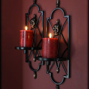 May include: Two black metal wall sconces with red pillar candles. Each sconce features a skull design and a decorative, geometric frame. The candles are lit, casting a warm glow. The sconces are mounted on a dark red wall.