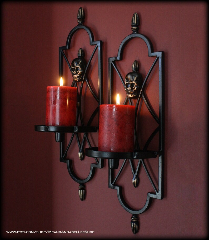 May include: Two black metal wall sconces with red pillar candles. Each sconce features a skull design and a decorative, geometric frame. The candles are lit, casting a warm glow. The sconces are mounted on a dark red wall.