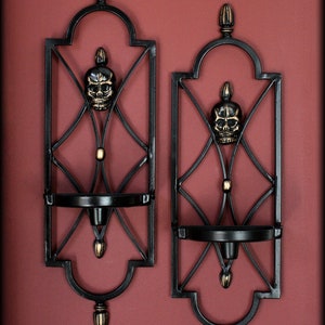 May include: Two black metal wall sconces with a skull design. Each sconce has a decorative frame with a skull in the center and a small shelf for a candle. The sconces are finished with gold accents.