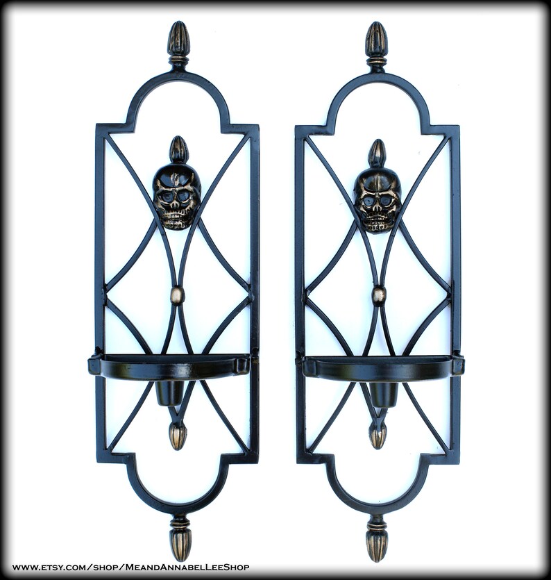 May include: Pair of black metal wall sconces with a skull design. Each sconce features a decorative frame with a skull in the center, a candle holder, and ornate detailing. The sconces are designed for wall mounting and add a gothic touch.