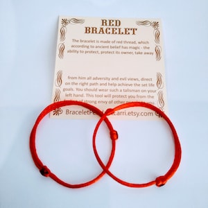 May include: Two bright red bracelets made of thread, with a card that reads "RED BRACELET". The card describes the bracelet's protective qualities. The bracelets are knotted and circular, displayed on a white surface.