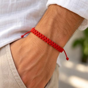 May include: A bright red woven cord bracelet featuring a repeating zigzag knot pattern. The bracelet is designed with adjustable pull strings on each side, ending in small knotted tips, allowing for a flexible fit around the wrist.
