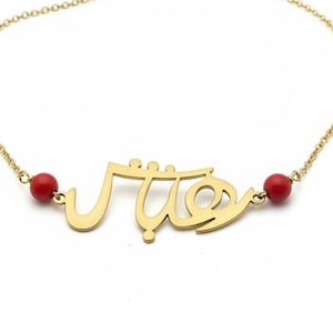Könnte beinhalten: Eine goldfarbene Halskette mit arabischem Schriftzug-Anhänger und zwei roten Perlen. Die Kette befindet sich auf weißem Hintergrund. Der Anhänger ist der Blickfang, mit der Schrift in einem stilisierten Schriftbild. Die roten Perlen setzen Farbakzente.