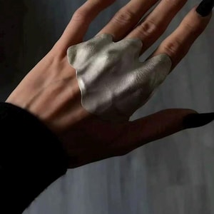 May include: A silver-colored ring with an organic, abstract design. The ring has a textured surface and a unique, flowing shape. The ring is worn on a finger with long, black fingernails. The ring is a piece of jewelry.