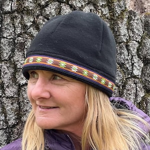 May include: A black beanie with a decorative band featuring a geometric pattern in shades of green, brown, and purple. The hat is worn by a person with blonde hair and a purple jacket, set against a textured tree trunk background.