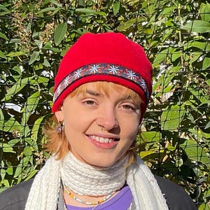 May include: A person wearing a red fleece hat with a black and white patterned band. The hat is pulled down over their forehead and they are smiling.