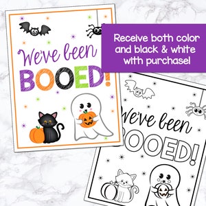 May include: A printable Halloween sign with the text "We've been BOOED!" in black, orange, and purple. The sign features a ghost holding a pumpkin, a black cat, and a spider. A black and white version of the sign is also included.