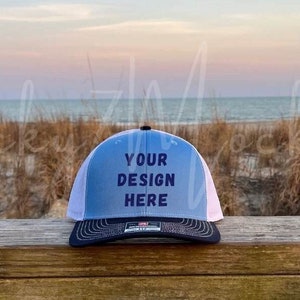 May include: A blue and white trucker hat with the text "YOUR DESIGN HERE" in navy blue. The hat has a black brim and is sitting on a wooden surface with a beach scene in the background.