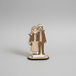 Puede incluir: Figurita de madera de una pareja, estilo dibujado a mano. La mujer lleva un vestido a rayas y una chaqueta, con un bolso. El hombre lleva un traje y una gorra. La base dice "Anna & Filippo".