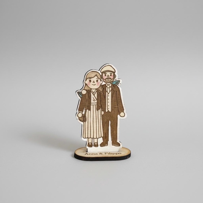 Puede incluir: Figurita de madera de una pareja, estilo dibujado a mano. La mujer lleva un vestido a rayas y una chaqueta, con un bolso. El hombre lleva un traje y una gorra. La base dice "Anna & Filippo".