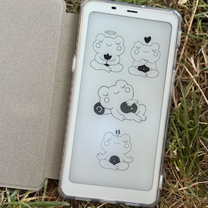 May include: A white e-reader with a clear protective case, displaying four cartoon frog illustrations in black. The e-reader is open, revealing the screen. A light gray cover is visible to the left. The e-reader is resting on grass.